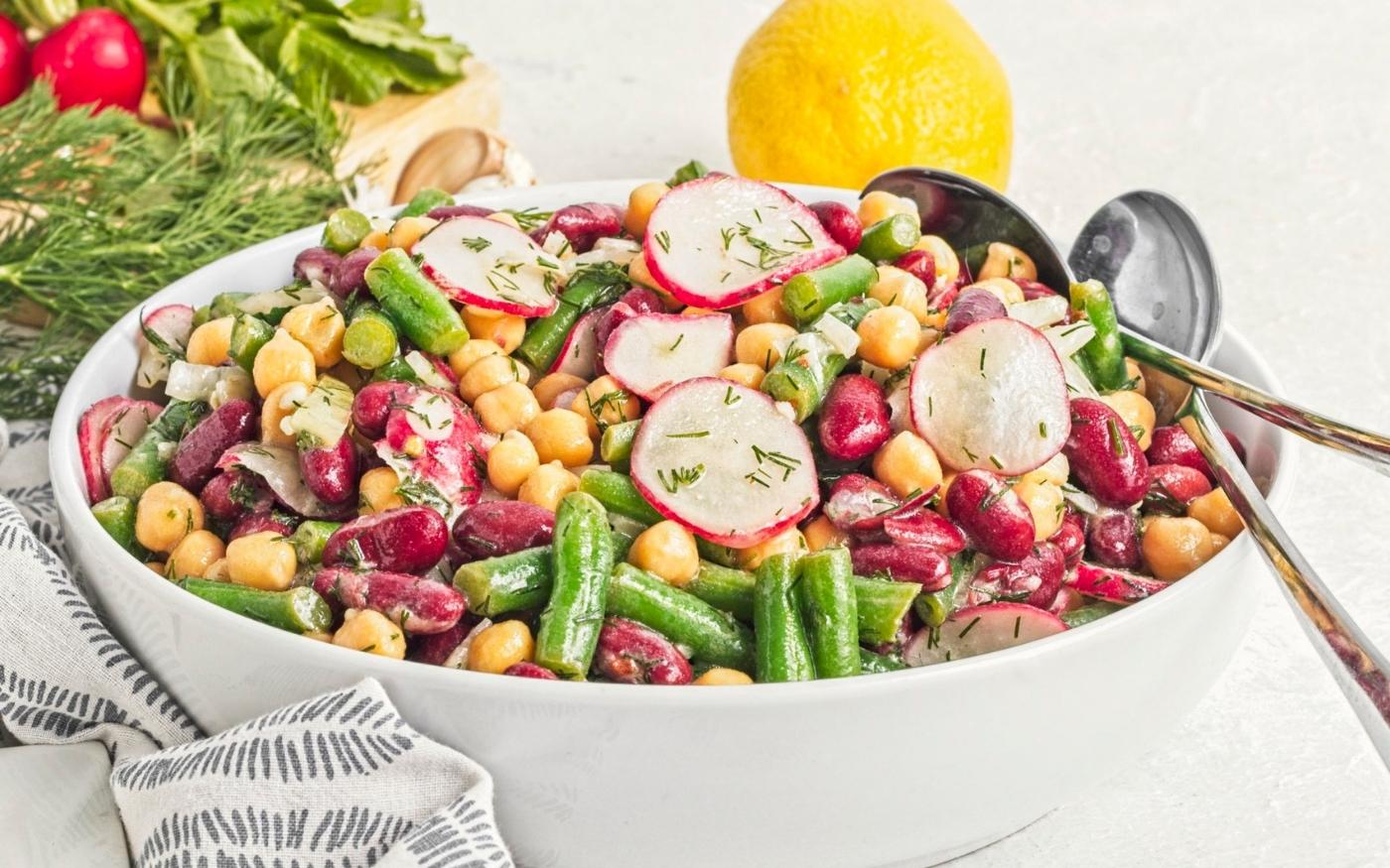 A bowl of mixed bean salad with chickpeas, kidney beans, green beans, sliced radishes, and herbs, with serving spoons and a lemon in the background.