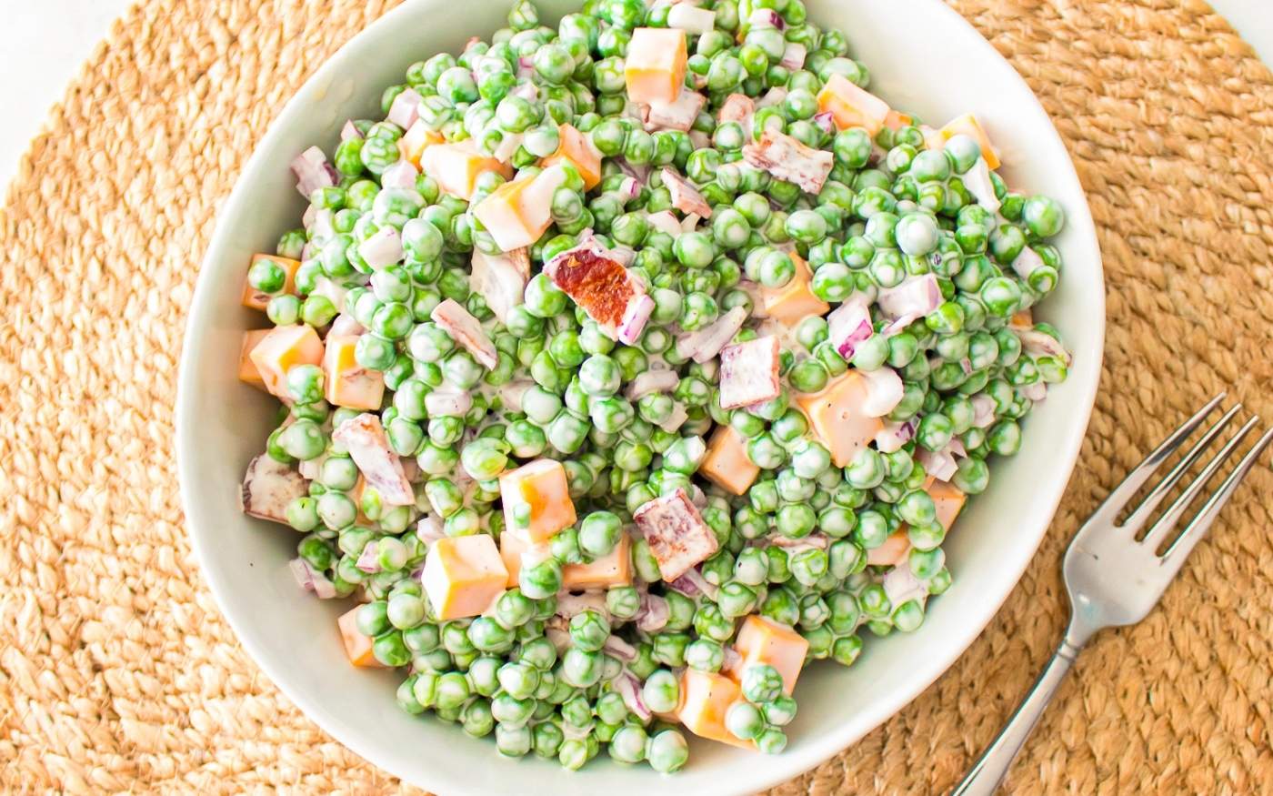 A white bowl filled with creamy pea salad containing green peas, cubed cheese, bacon bits, and diced red onion, placed on a woven placemat next to a fork.