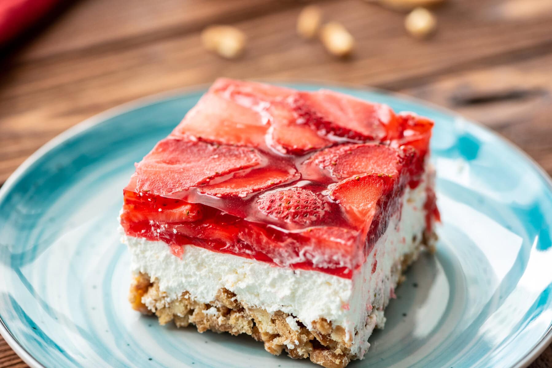 A square slice of dessert with a crumbly base, creamy middle layer, and strawberry topping in clear gelatin, served on a blue plate.
