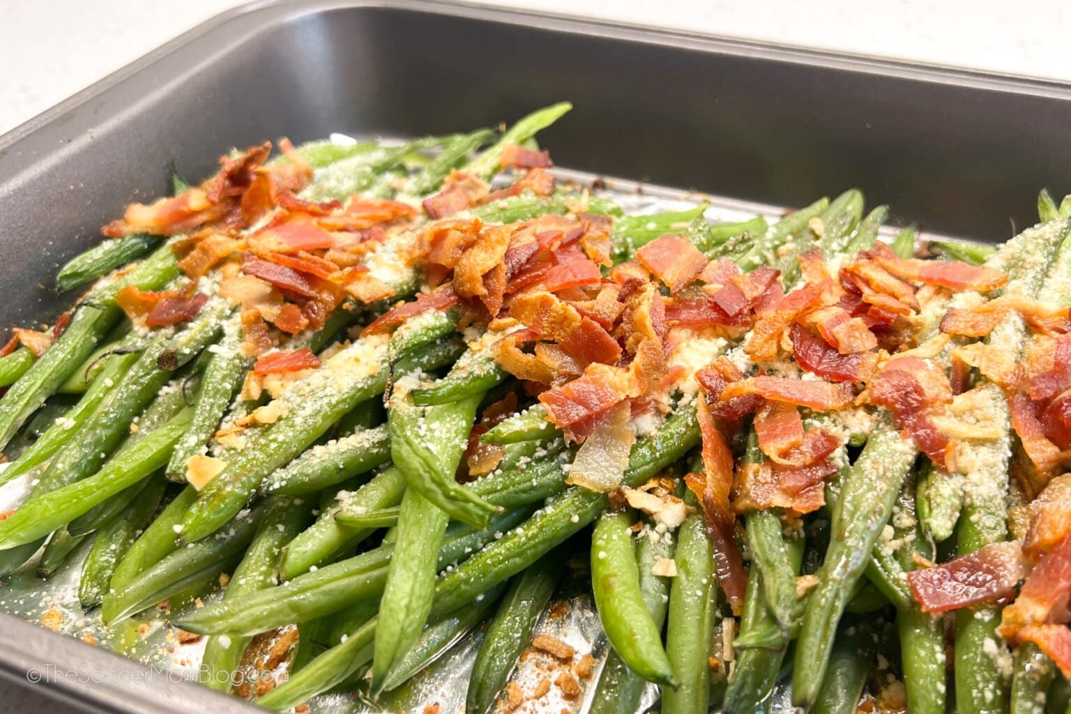 A baking dish filled with cooked green beans topped with chopped bacon and grated cheese.