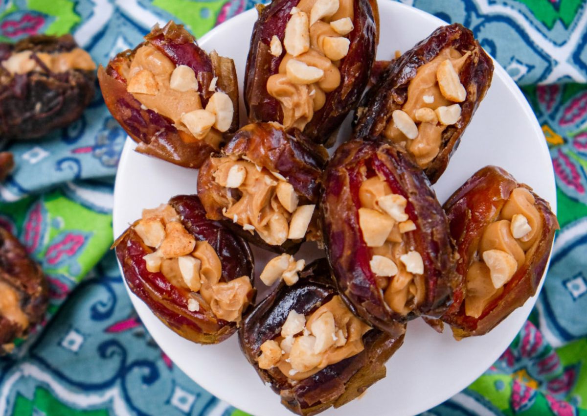 A white plate holds dates stuffed with peanut butter and chopped nuts, set on a patterned blue and green fabric background.