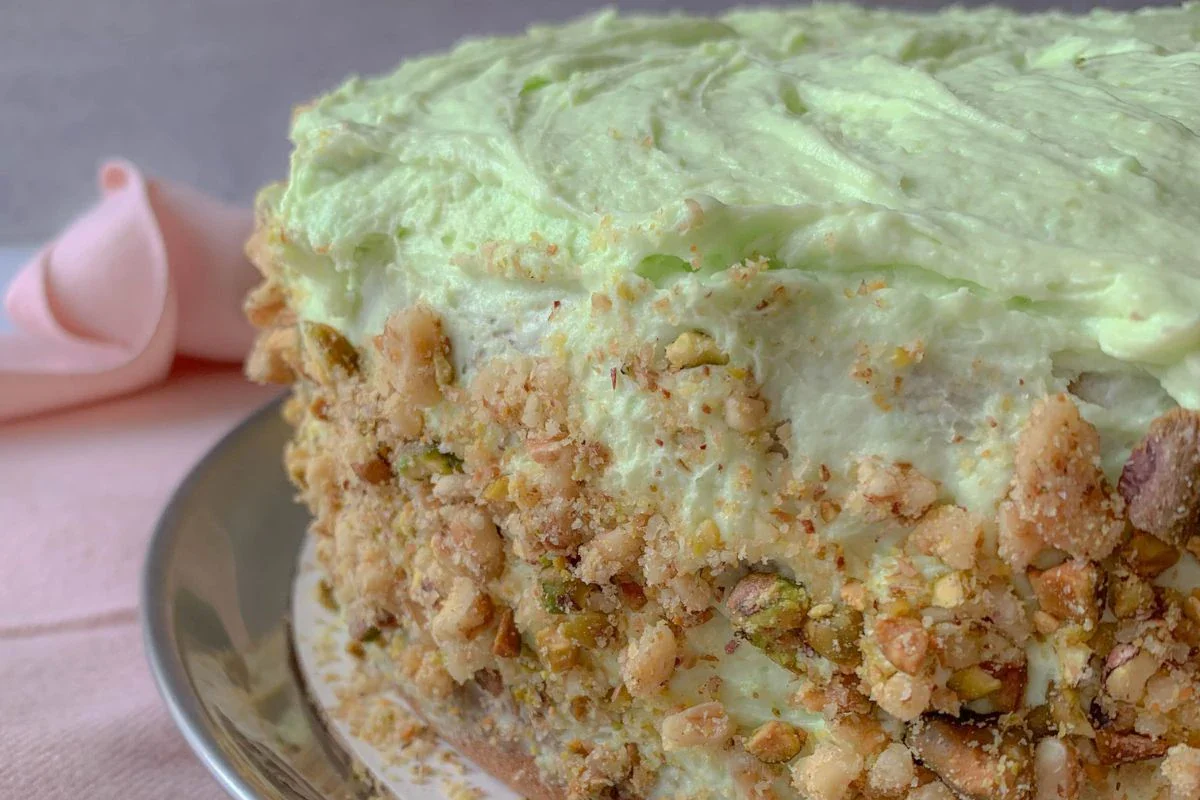 A slice of cake with pale green frosting and crushed nuts on the sides, served on a plate with a pink napkin in the background.