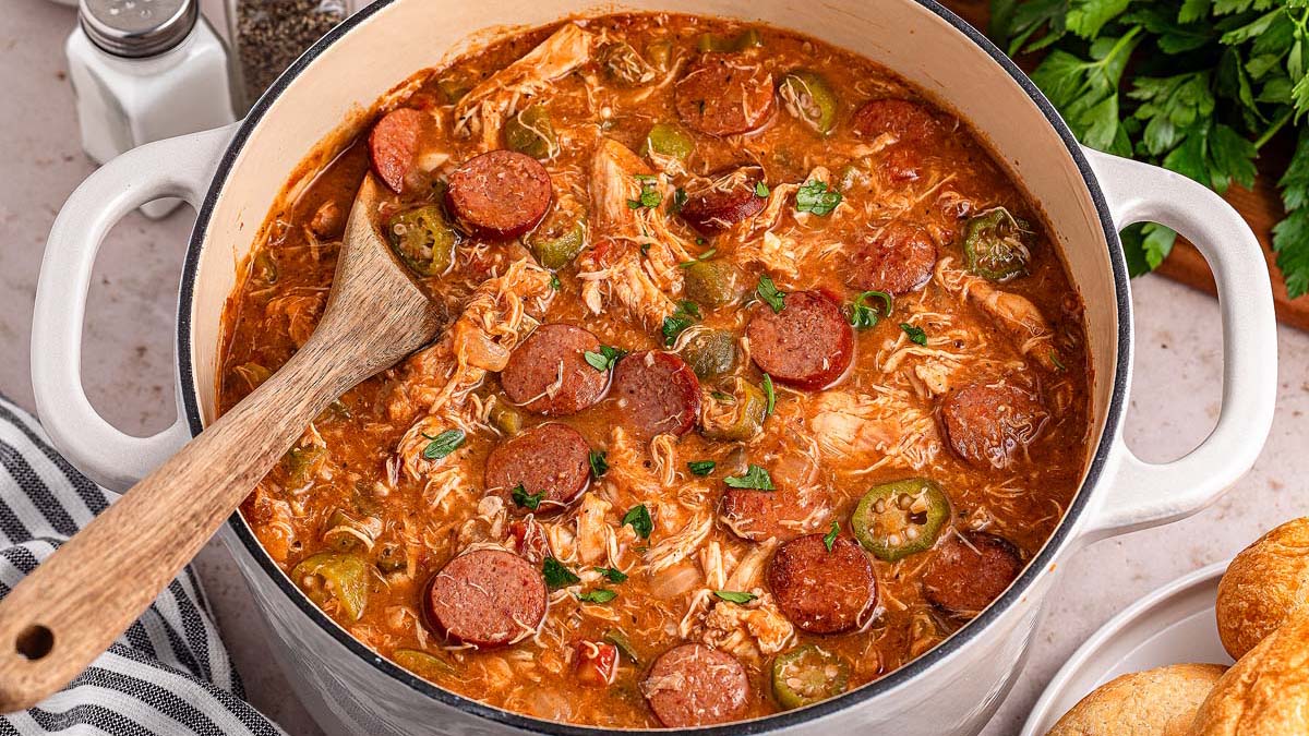A pot of gumbo with sliced sausage, shredded chicken, and vegetables in a rich broth, with a wooden spoon resting inside the pot.