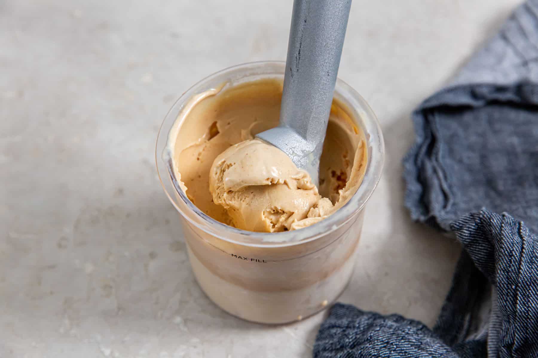 A metal ice cream scoop is in a container of light brown ice cream, with a dark blue cloth on a light countertop nearby.