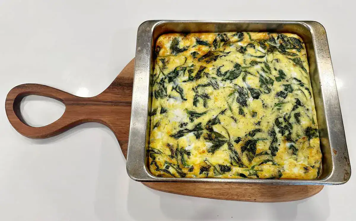 A baked spinach and egg casserole in a square metal pan on a wooden cutting board, viewed from above.