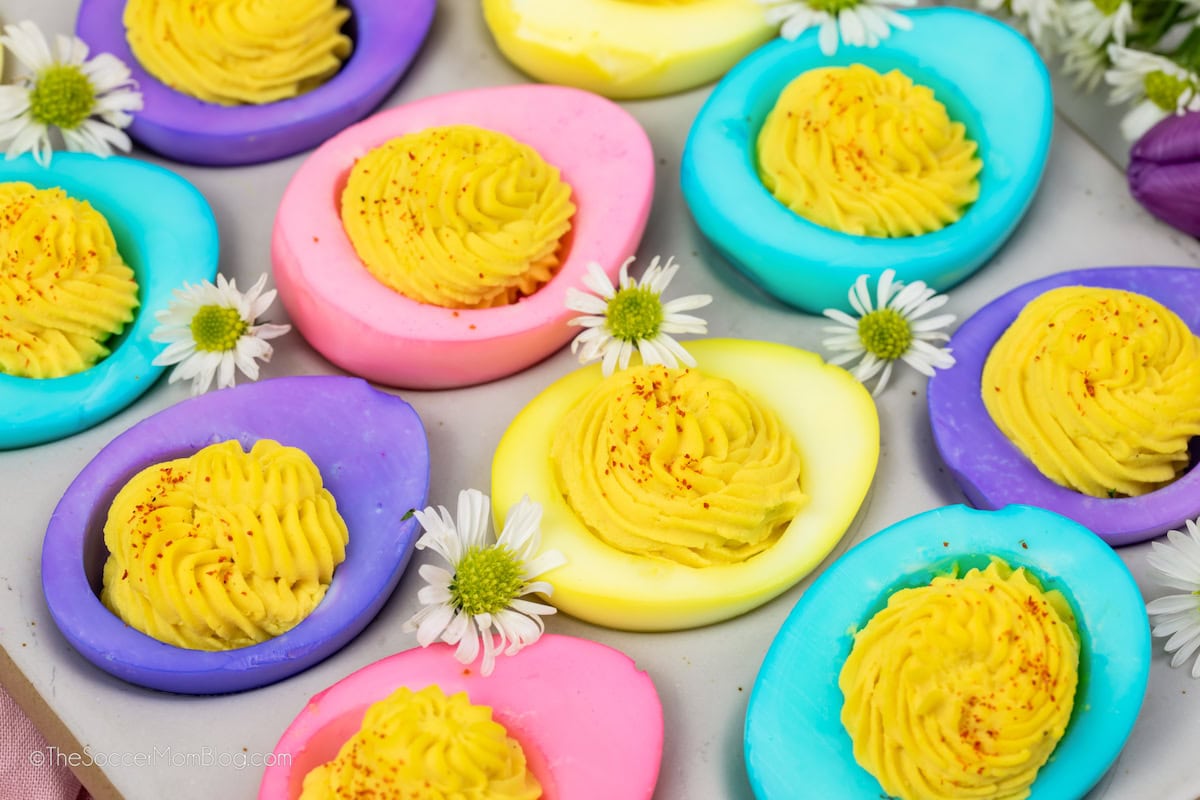 Colorful deviled eggs with swirled yellow filling are arranged on a surface with small white daisy flowers scattered between them.
