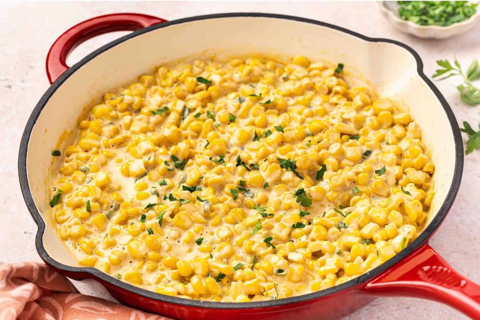 Creamed corn garnished with chopped herbs in a red enameled cast iron skillet, placed on a light countertop with a cloth and herbs nearby.
