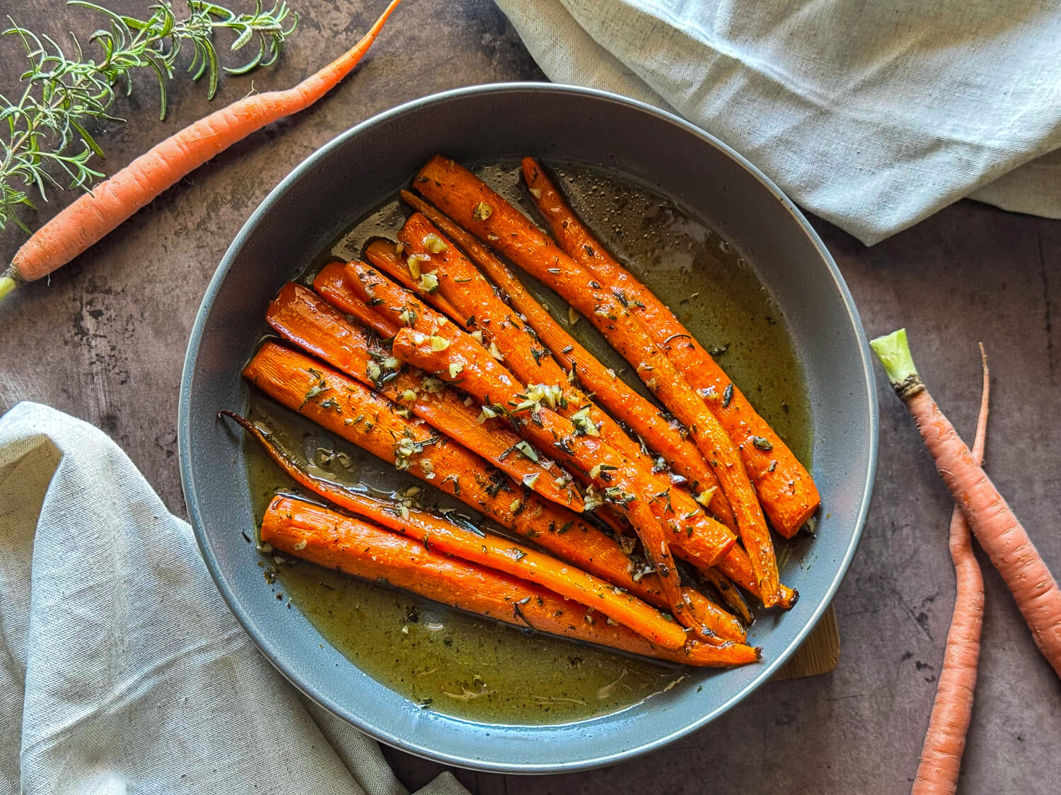 Cooked carrots in a round pan with herbs and garlic in a sauce, surrounded by raw carrots, rosemary, and beige cloths on a wooden surface.