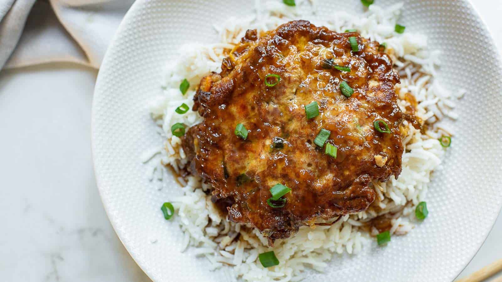 A plate of white rice topped with a glazed meat patty and garnished with chopped green onions.