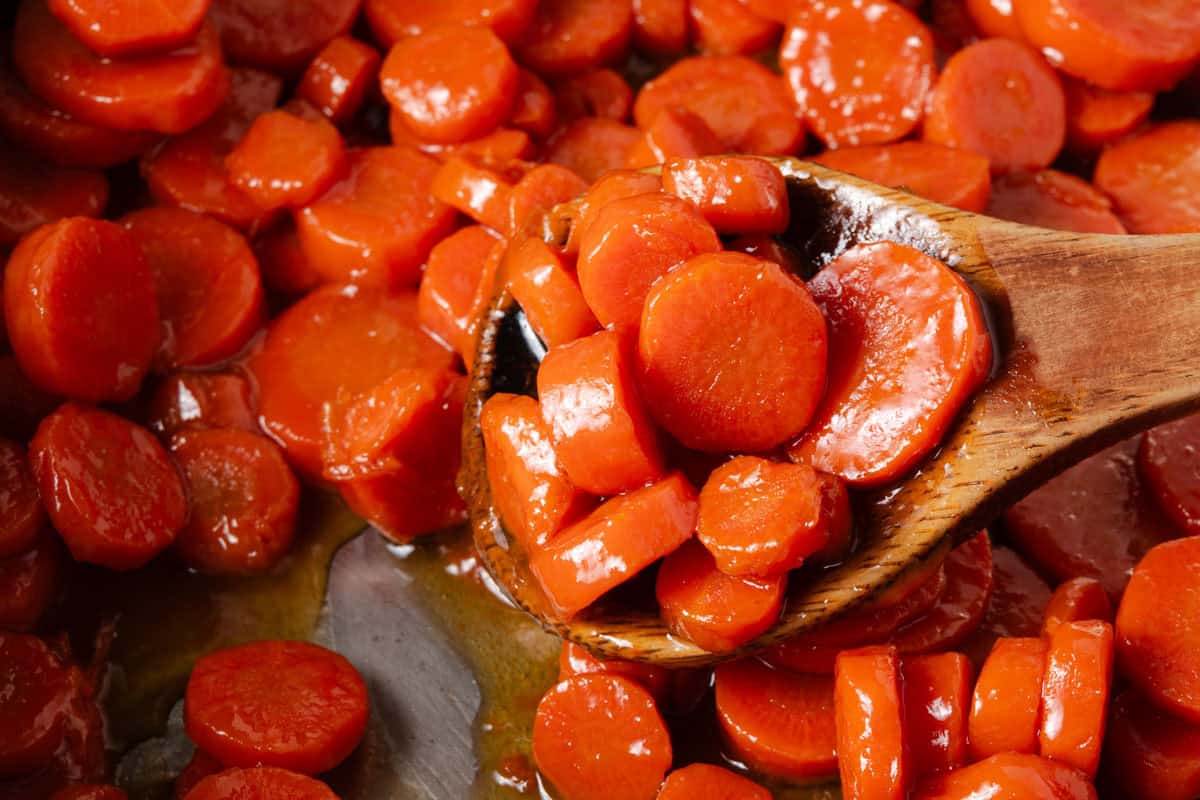 A wooden spoon scoops glazed carrot slices from a pan filled with more glazed carrots.
