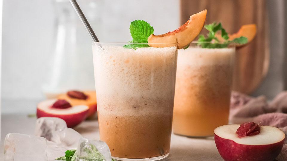 Two glasses of peach smoothie with frothy tops, garnished with peach slices and mint leaves, sit on a table with ice cubes and halved peaches nearby.
