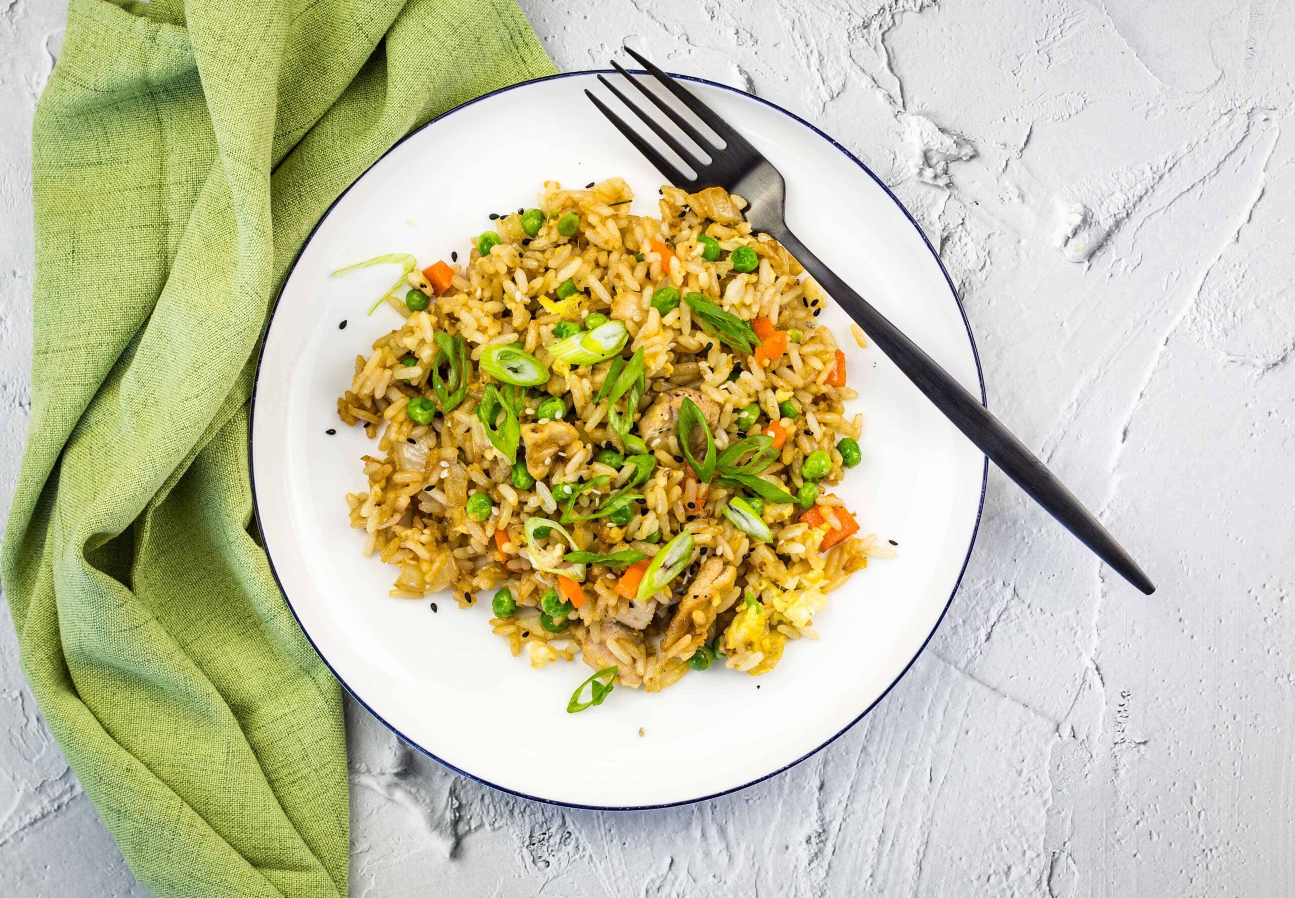 A plate of vegetable fried rice with carrots, peas, and green onions, placed next to a black fork and a green napkin on a light textured surface.