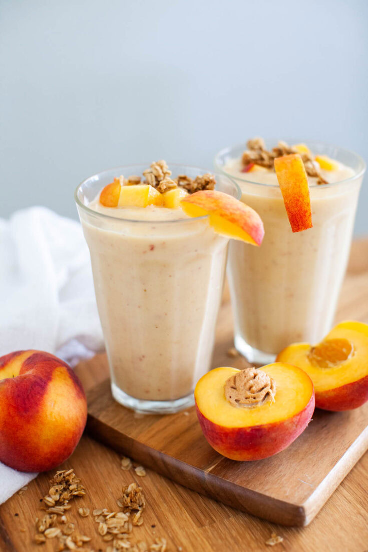 Two glasses of peach smoothie topped with peach slices and granola sit on a wooden board next to a halved peach and scattered granola.