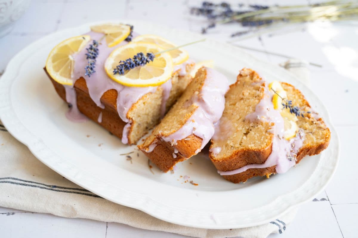 A loaf cake with light purple icing is topped with lemon slices and lavender sprigs; two slices are cut and arranged on a white plate.