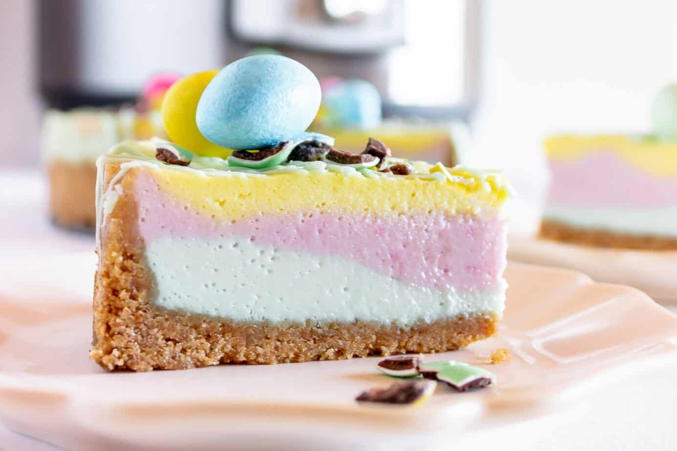 Slice of layered cheesecake with pastel pink, yellow, and white filling on a graham cracker crust, topped with candy eggs and chocolate pieces on a plate.