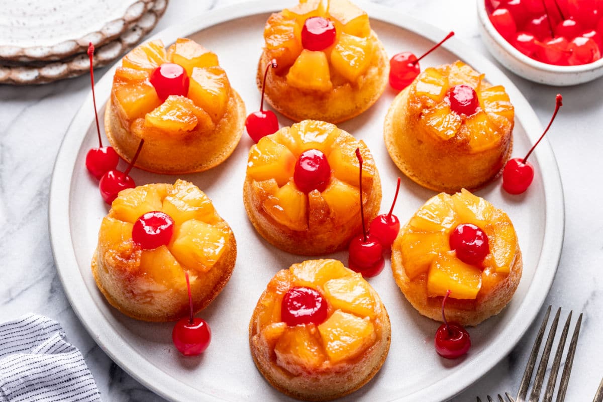 A plate of seven mini pineapple upside-down cakes, each topped with a cherry, with extra cherries in a bowl nearby.