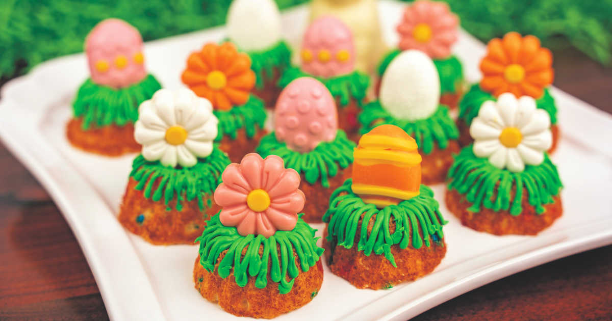Mini bundt cakes decorated with green icing, topped with pastel-colored candy Easter eggs and flowers, displayed on a white tray.