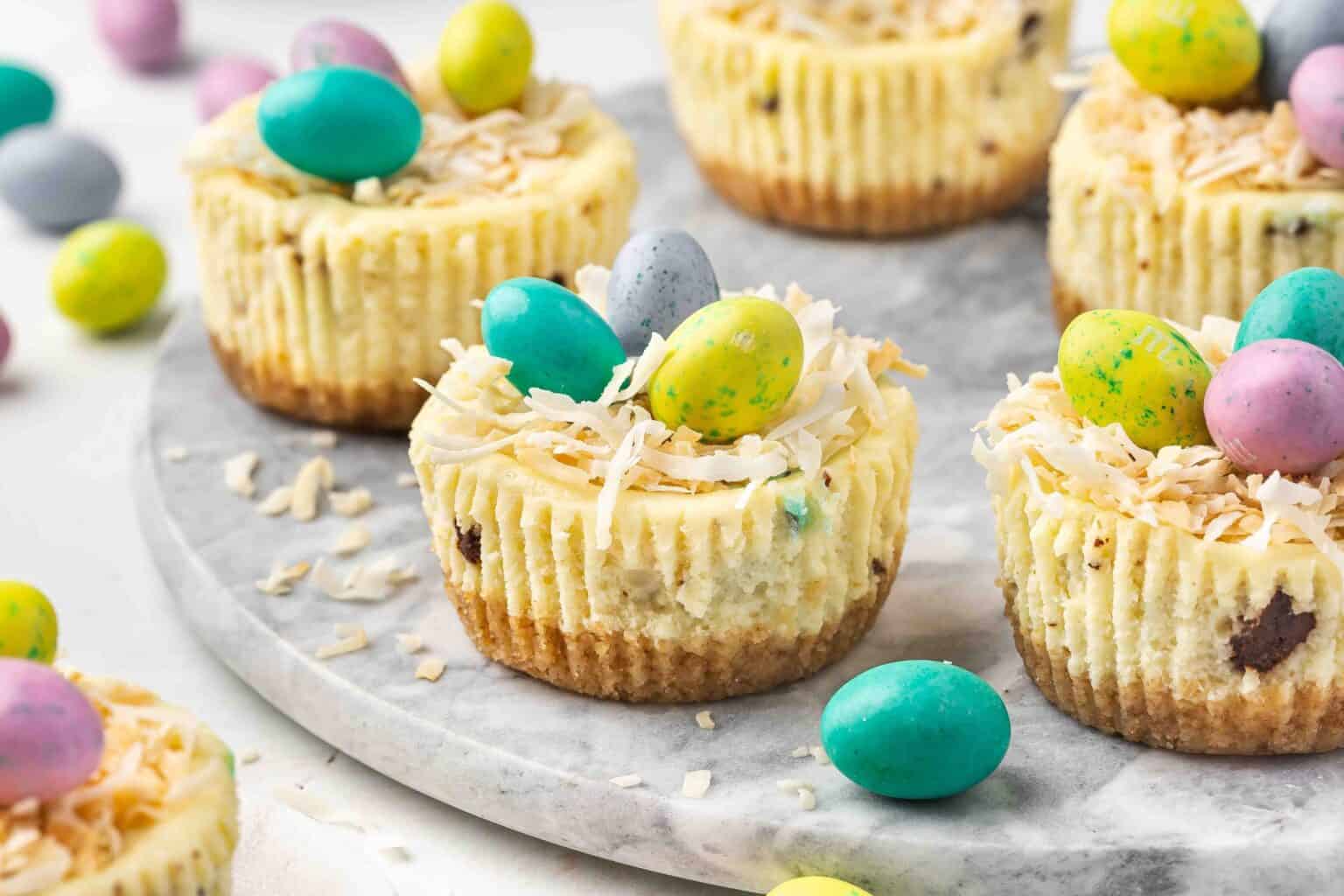 Mini cheesecakes topped with shredded coconut and colorful candy eggs are arranged on a marble tray with extra candy eggs scattered around.