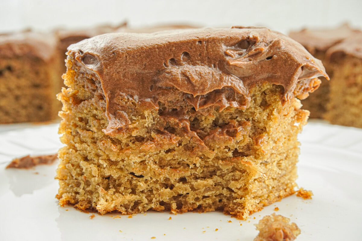 A close-up of a square piece of cake with light brown frosting on top, placed on a white plate.