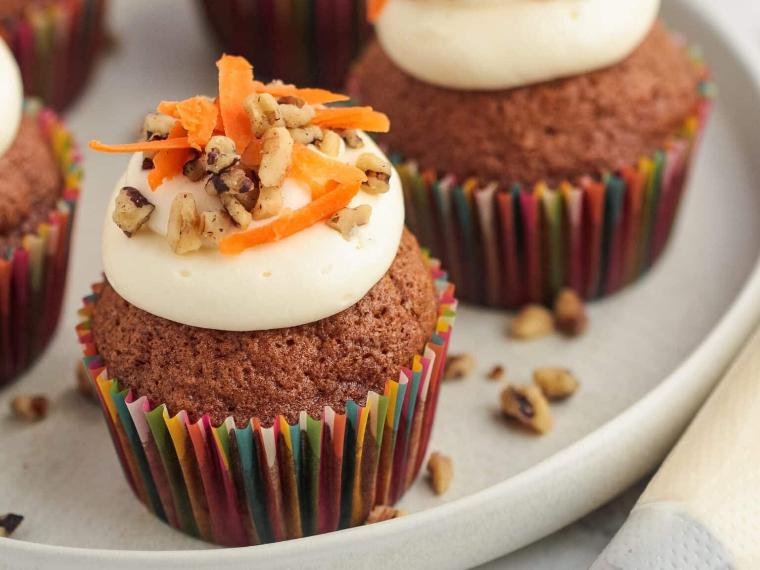 A carrot cupcake with cream cheese frosting, topped with chopped walnuts and shredded carrots, in a colorful paper liner on a plate.