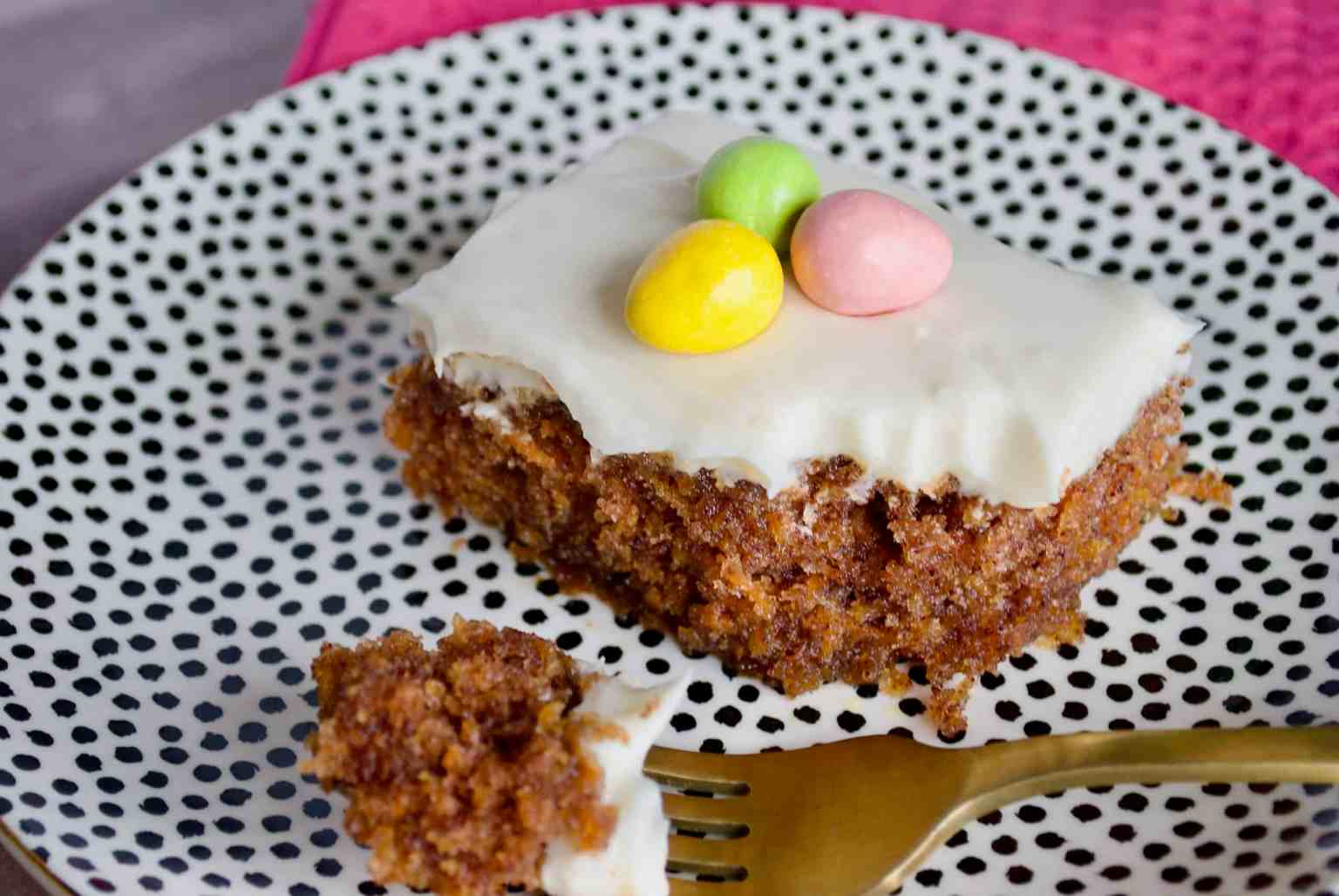 A slice of carrot cake with white icing and three colorful candy eggs on top, served on a dotted plate with a gold fork.
