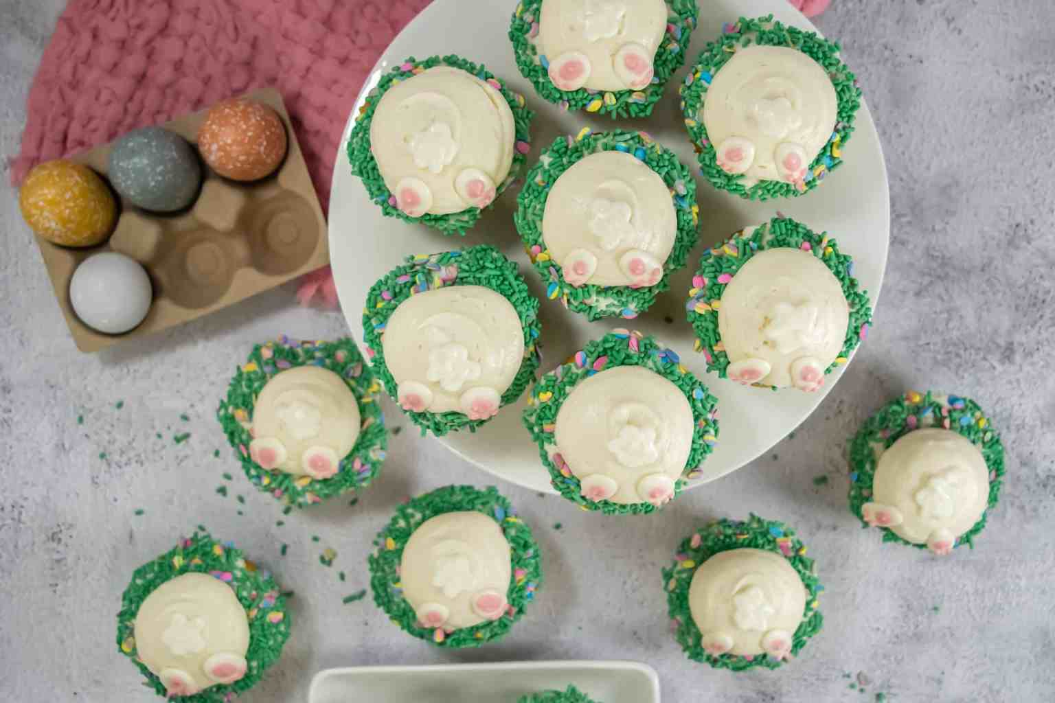 Cupcakes decorated with bunny bottoms and green sprinkles are arranged on a plate and table, next to a tray of colorful glittery eggs.