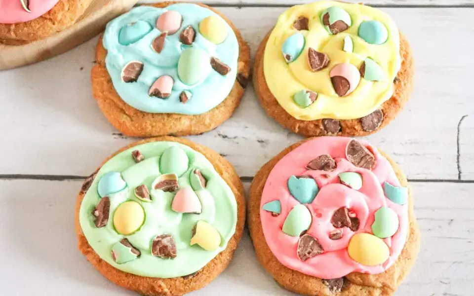 Four cookies on a white surface, each topped with colorful pastel icing and pieces of chocolate candy.
