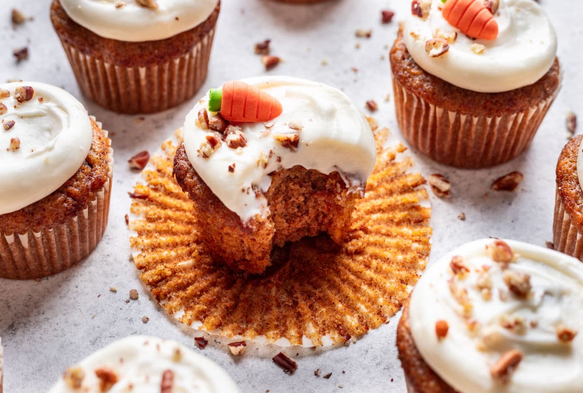 A group of carrot cupcakes with cream cheese frosting; one cupcake has a bite taken out and a small carrot-shaped decoration on top.