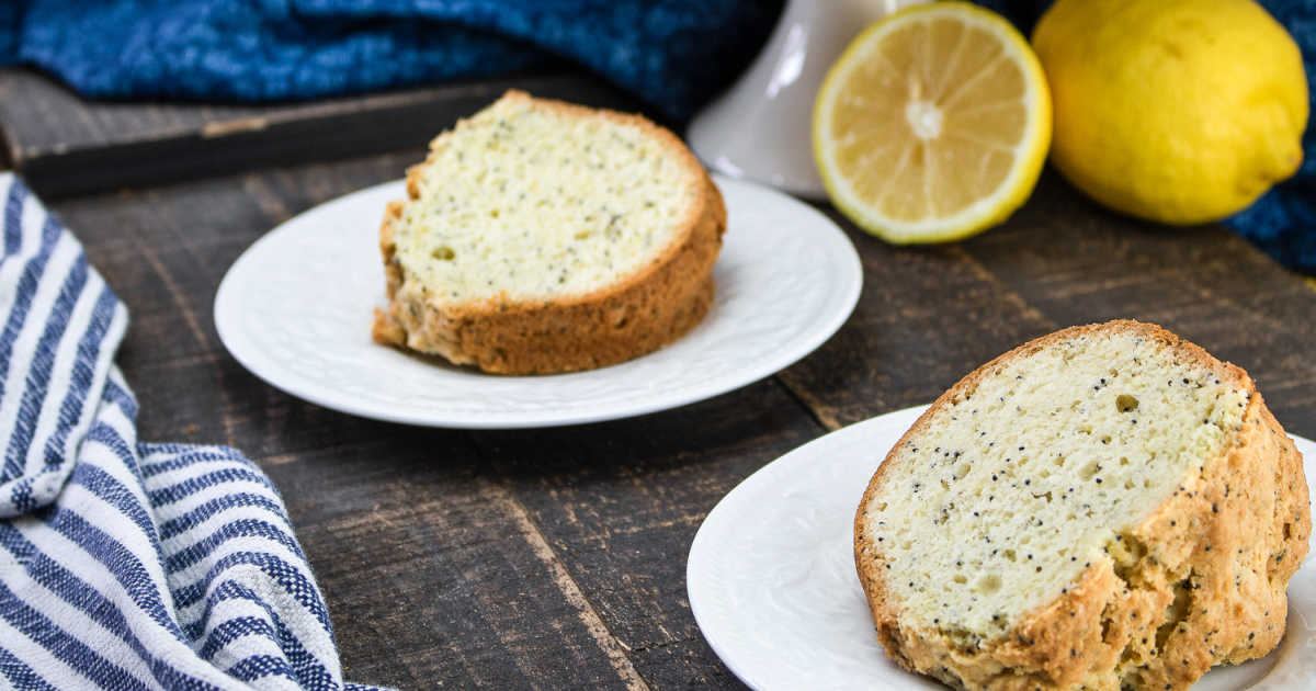 Two slices of poppy seed lemon cake on white plates sit on a wooden table, with a cut lemon and a striped cloth nearby.