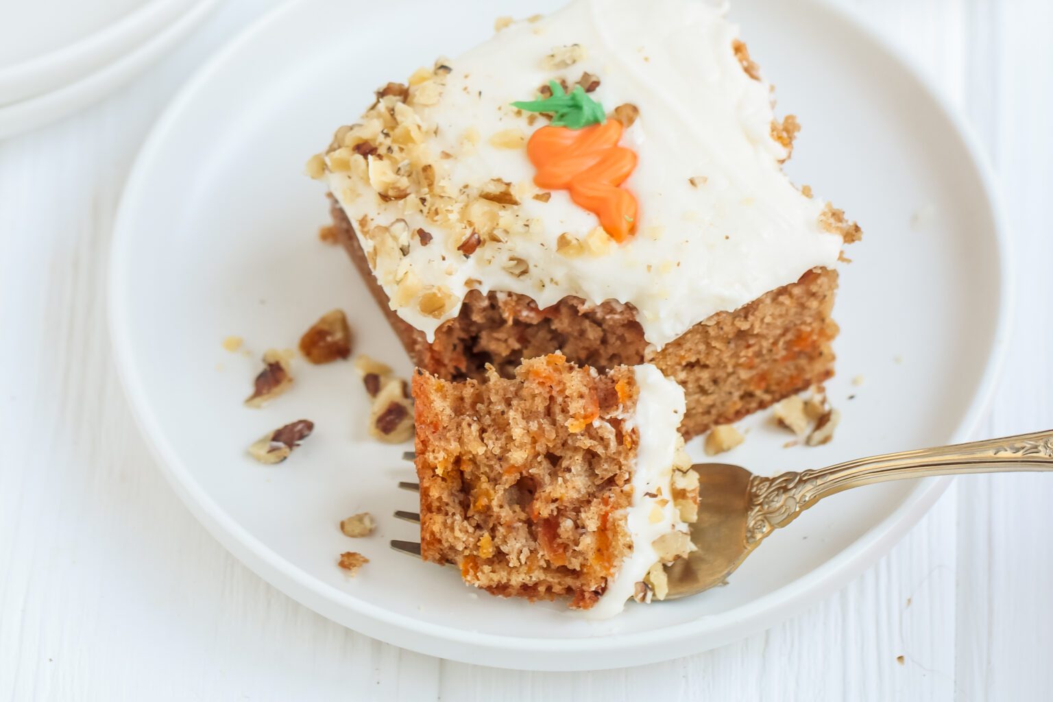 A piece of carrot cake with cream cheese frosting, chopped walnuts, and a small carrot decoration on top, served on a white plate with a fork holding a bite-sized piece.