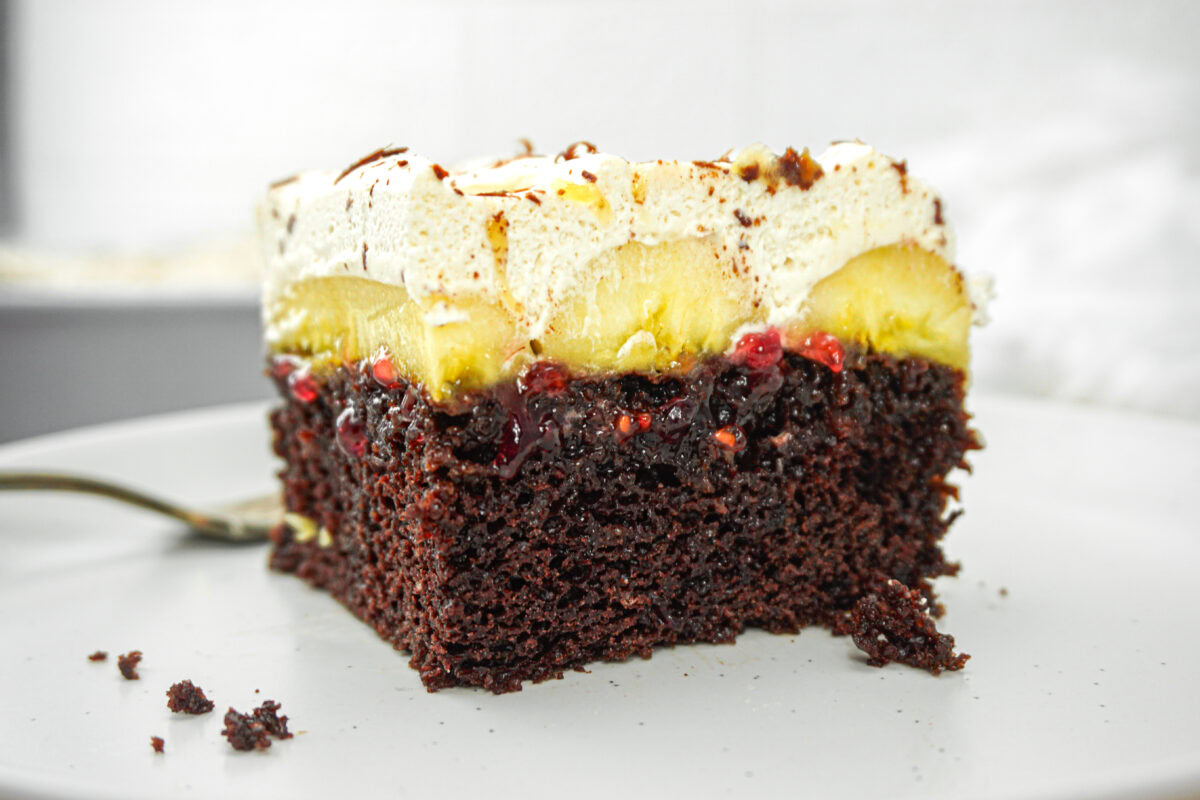 A slice of layered chocolate cake with cream, banana slices, fruit jam, and whipped topping on a white plate.