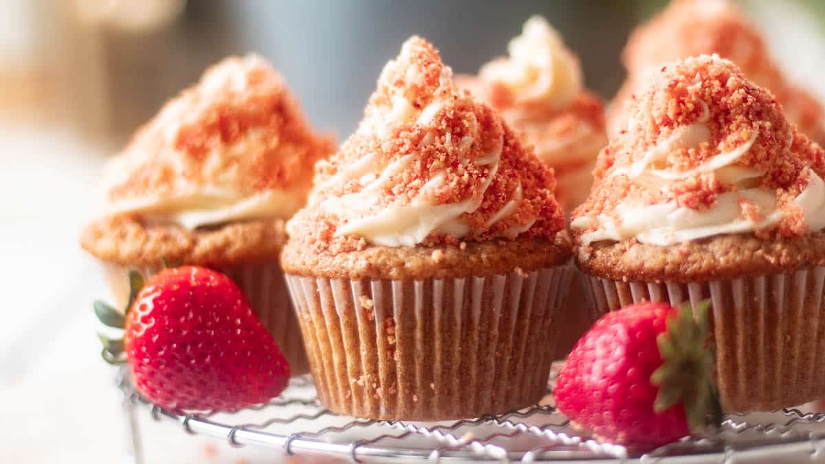 Three cupcakes with swirled frosting and crumb topping sit on a cooling rack beside two fresh strawberries.