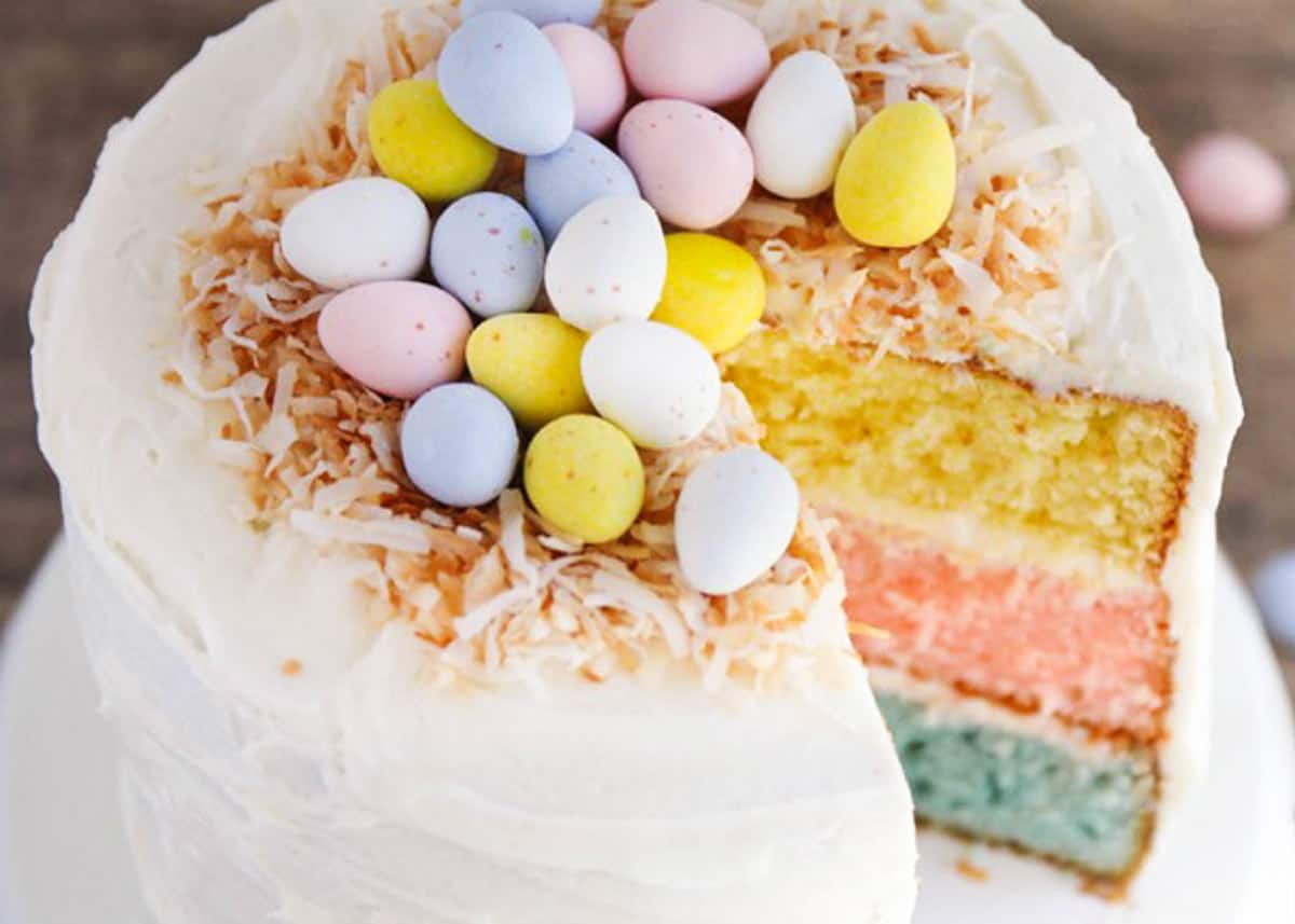 A three-layer cake with white frosting, topped with toasted coconut and pastel-colored candy eggs, with one slice missing.