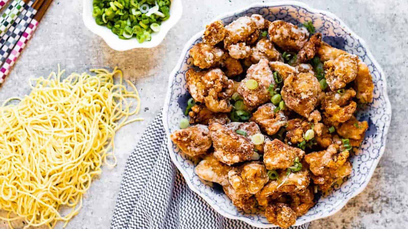 A bowl of crispy fried chicken pieces garnished with chopped green onions, with a side of uncooked noodles and a small bowl of sliced green onions.