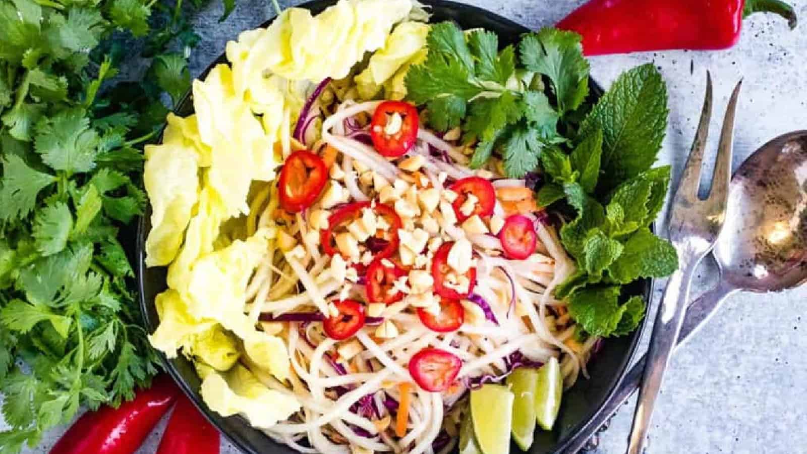 A bowl of Thai papaya salad topped with chopped peanuts and sliced red chili, surrounded by lettuce, lime wedges, fresh herbs, and silver utensils.