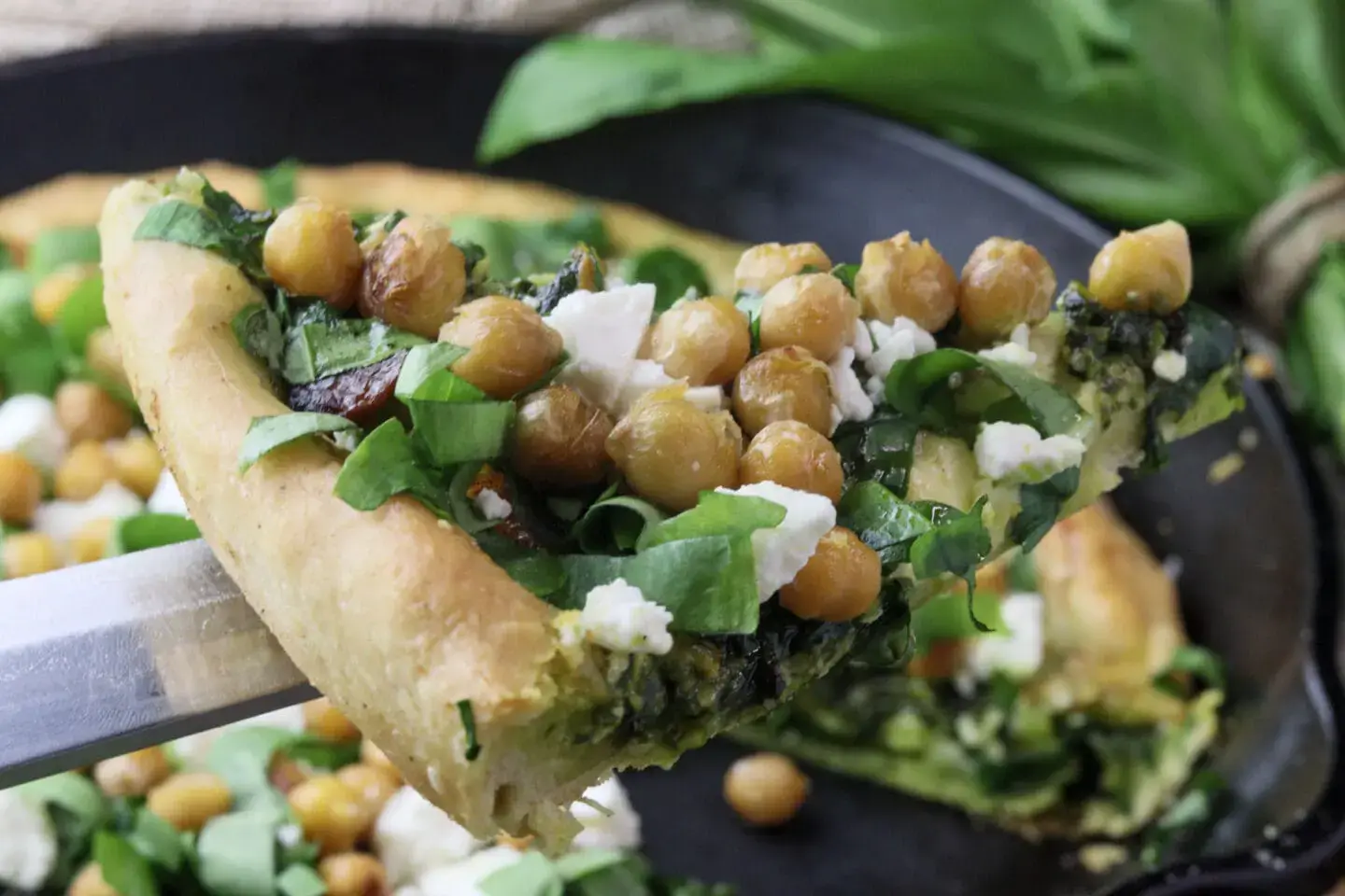 A slice of pizza topped with chickpeas, spinach, crumbled cheese, and fresh greens being lifted from a pan.