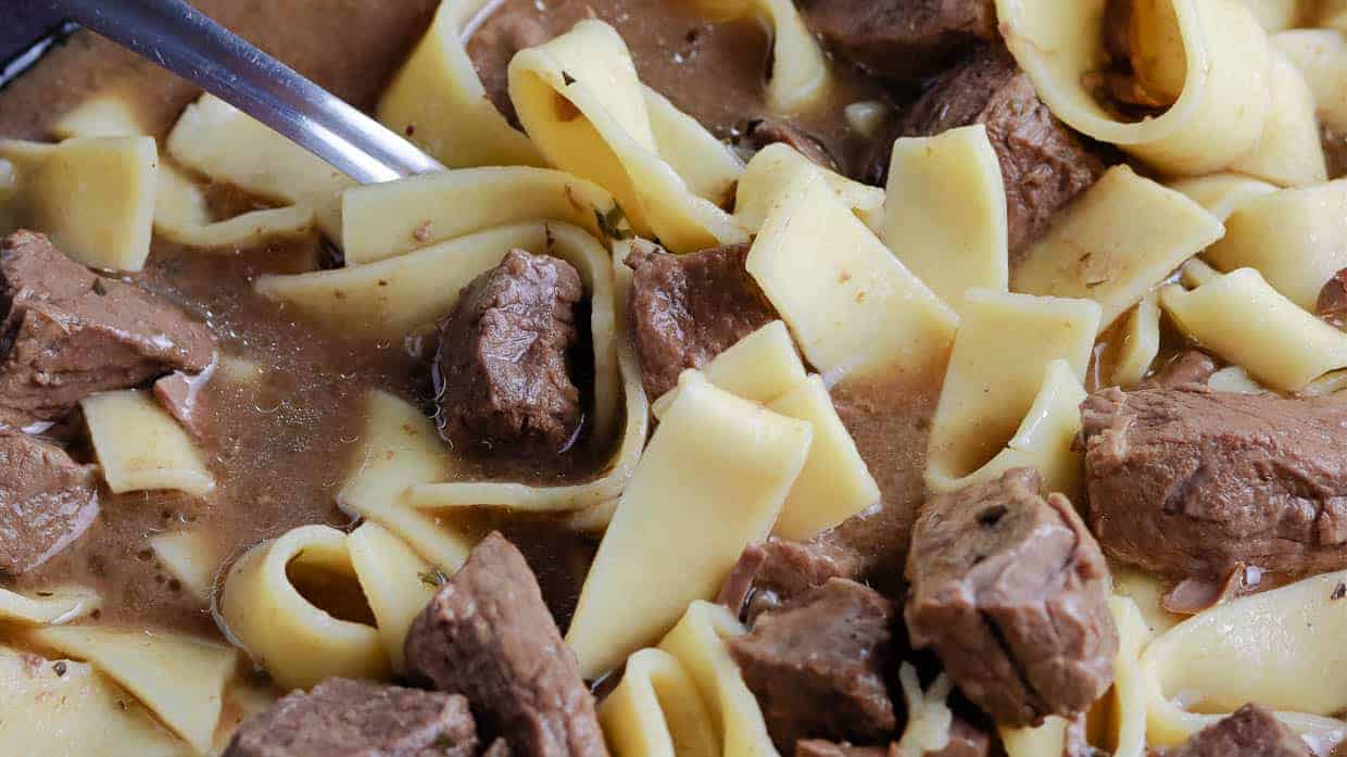 Wide egg noodles and chunks of beef in brown gravy with a spoon partially visible.