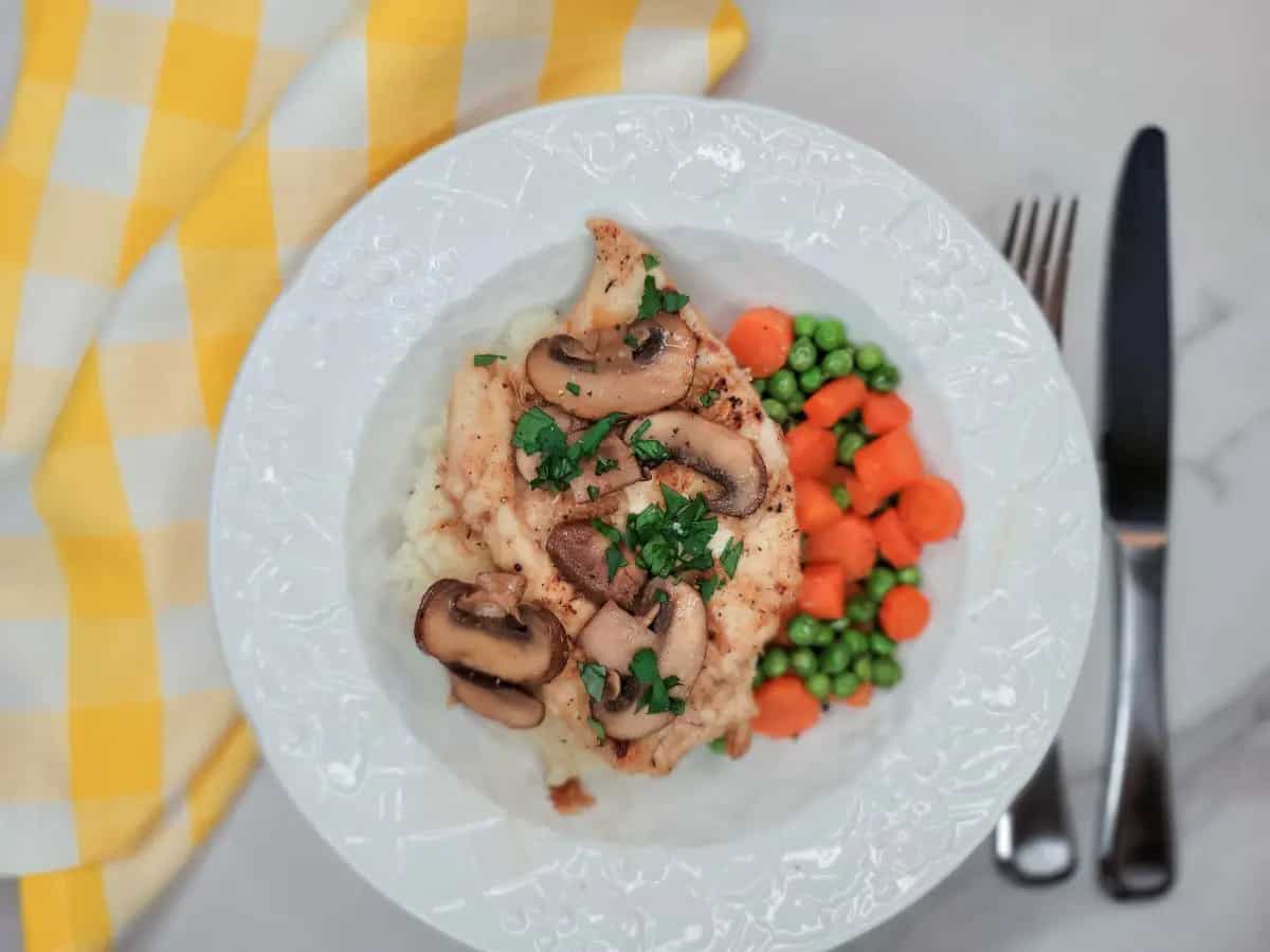 A plate with chicken topped with mushrooms and herbs, served with mashed potatoes, peas, and carrots; knife and fork on the side.