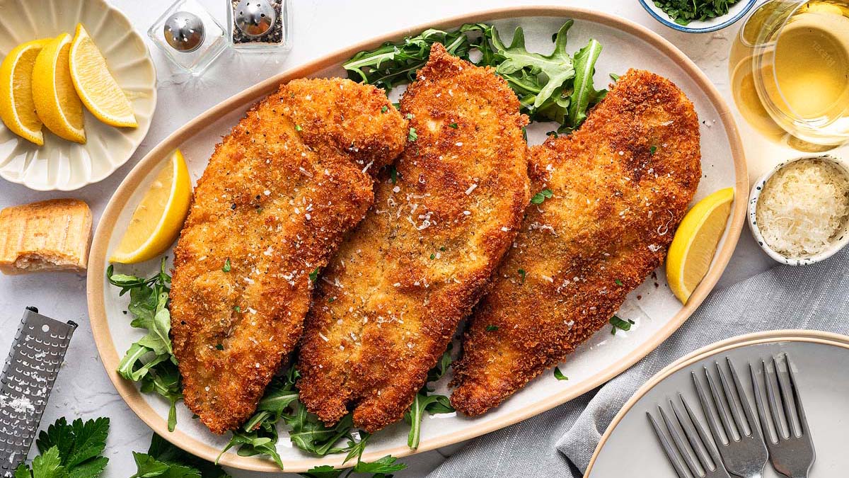 Three breaded and fried chicken cutlets on a bed of greens, topped with grated cheese, served with lemon wedges on a platter.