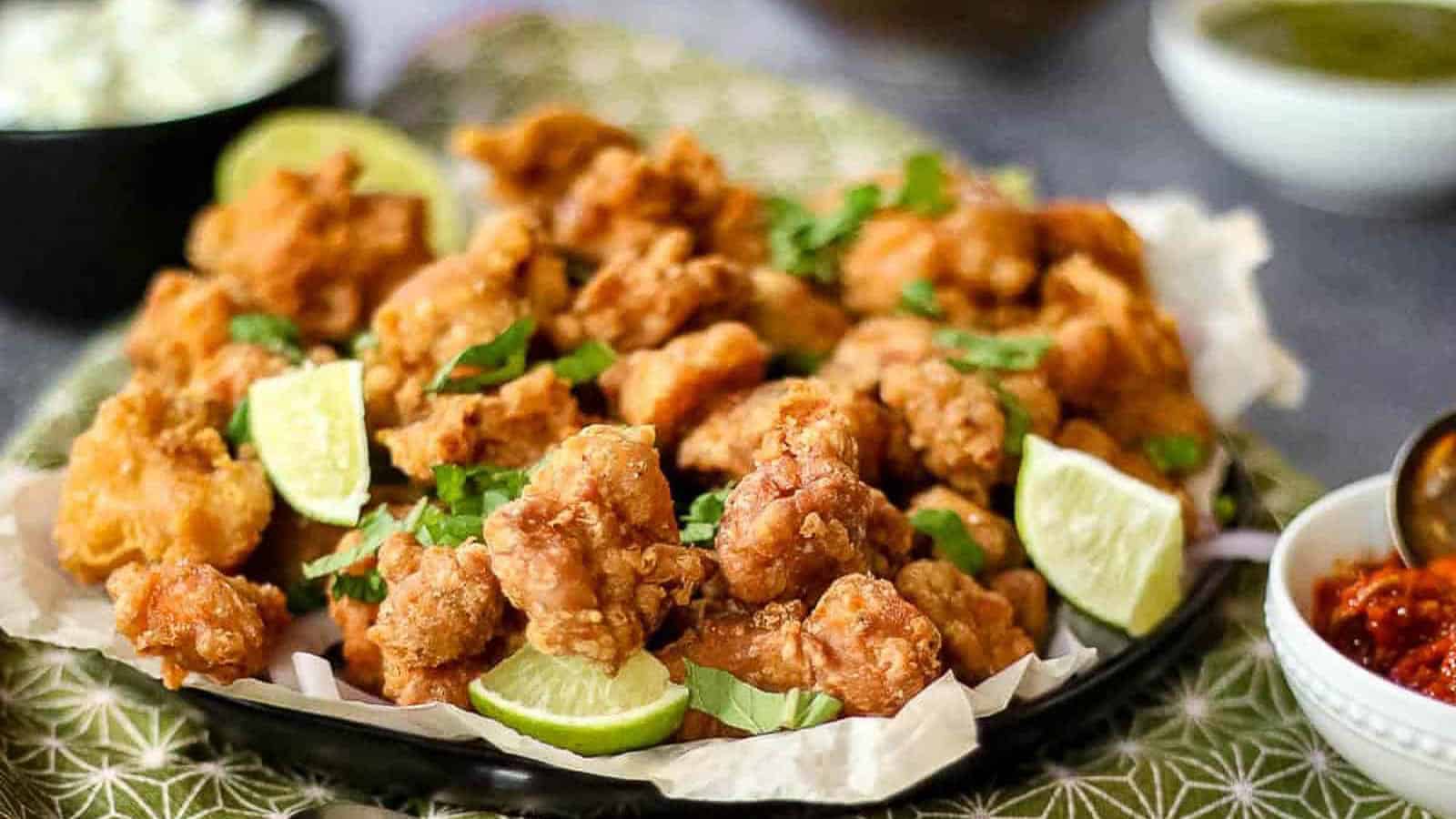 A plate of golden fried chicken pieces garnished with lime wedges and chopped herbs, served with sides in small bowls.