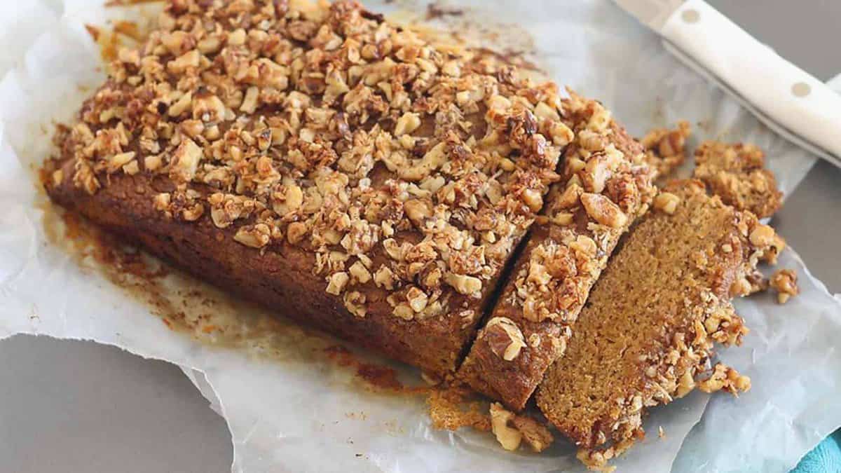 A loaf of nut-topped banana bread rests on parchment paper with two slices cut; a knife is placed nearby.
