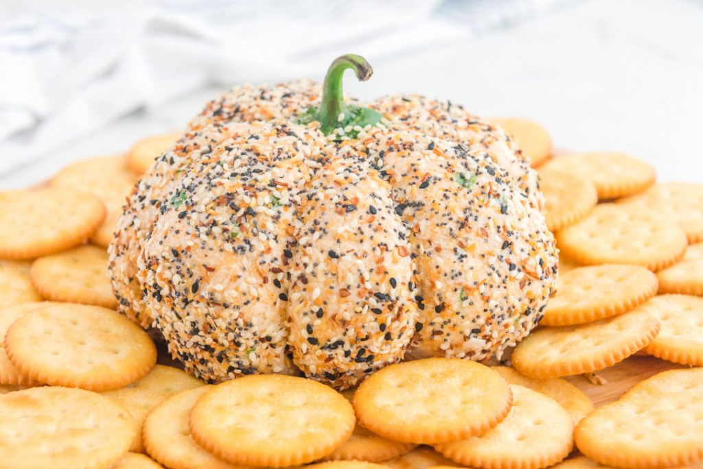 A cheese ball shaped like a pumpkin, coated with seasoning and topped with a green stem, surrounded by round crackers on a wooden board.