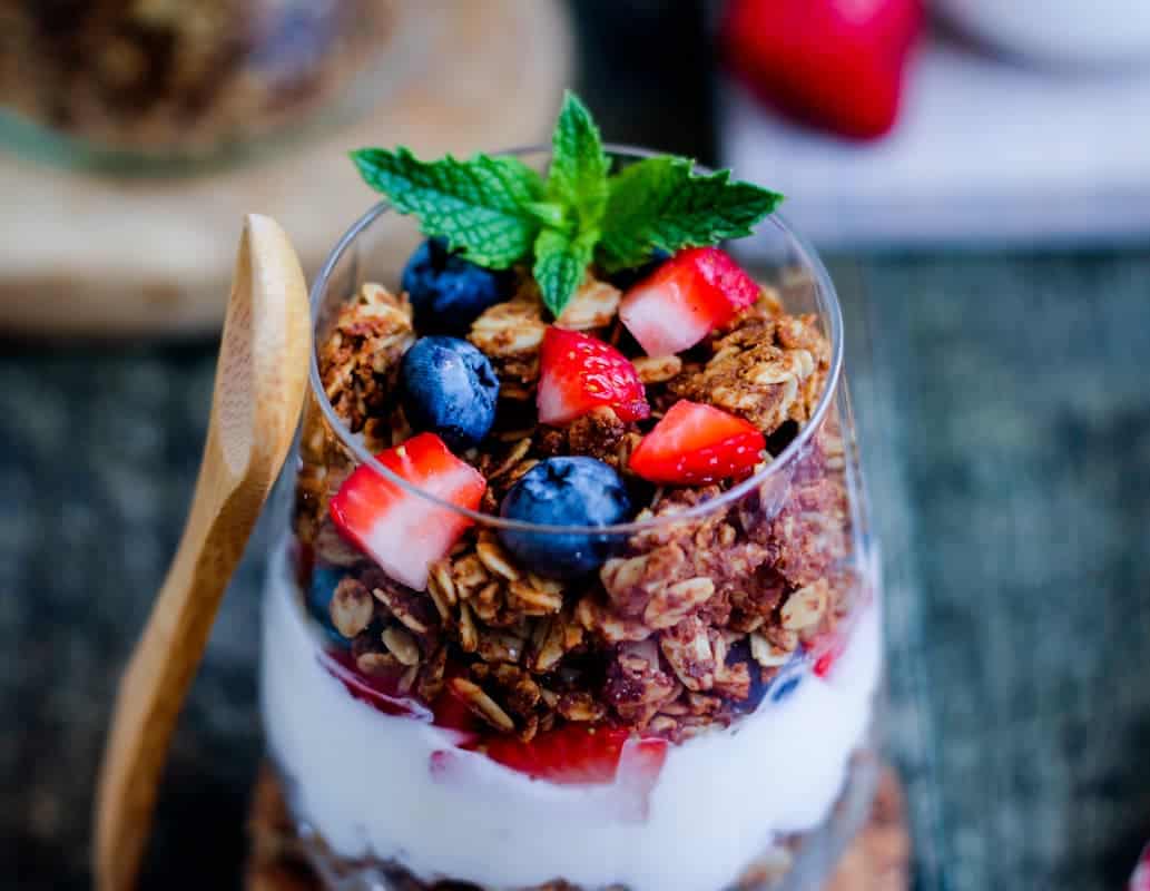 A glass cup filled with layers of yogurt, granola, strawberries, and blueberries, topped with fresh mint, sits beside a wooden spoon.