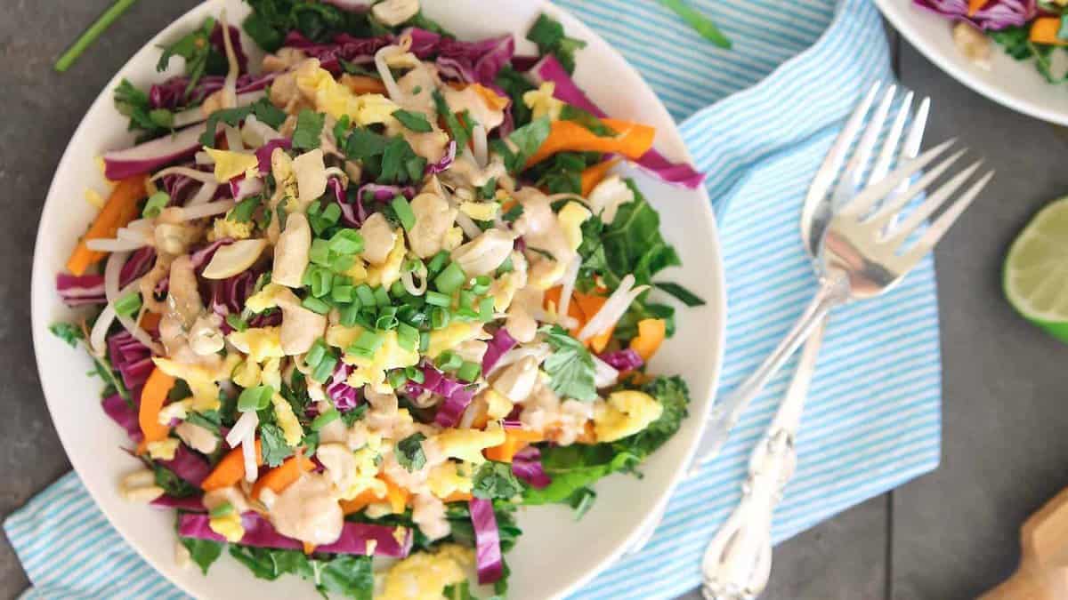 A bowl of colorful chopped salad with green onions, shredded carrots, cabbage, greens, and a creamy dressing, placed on a striped cloth with a fork beside it.