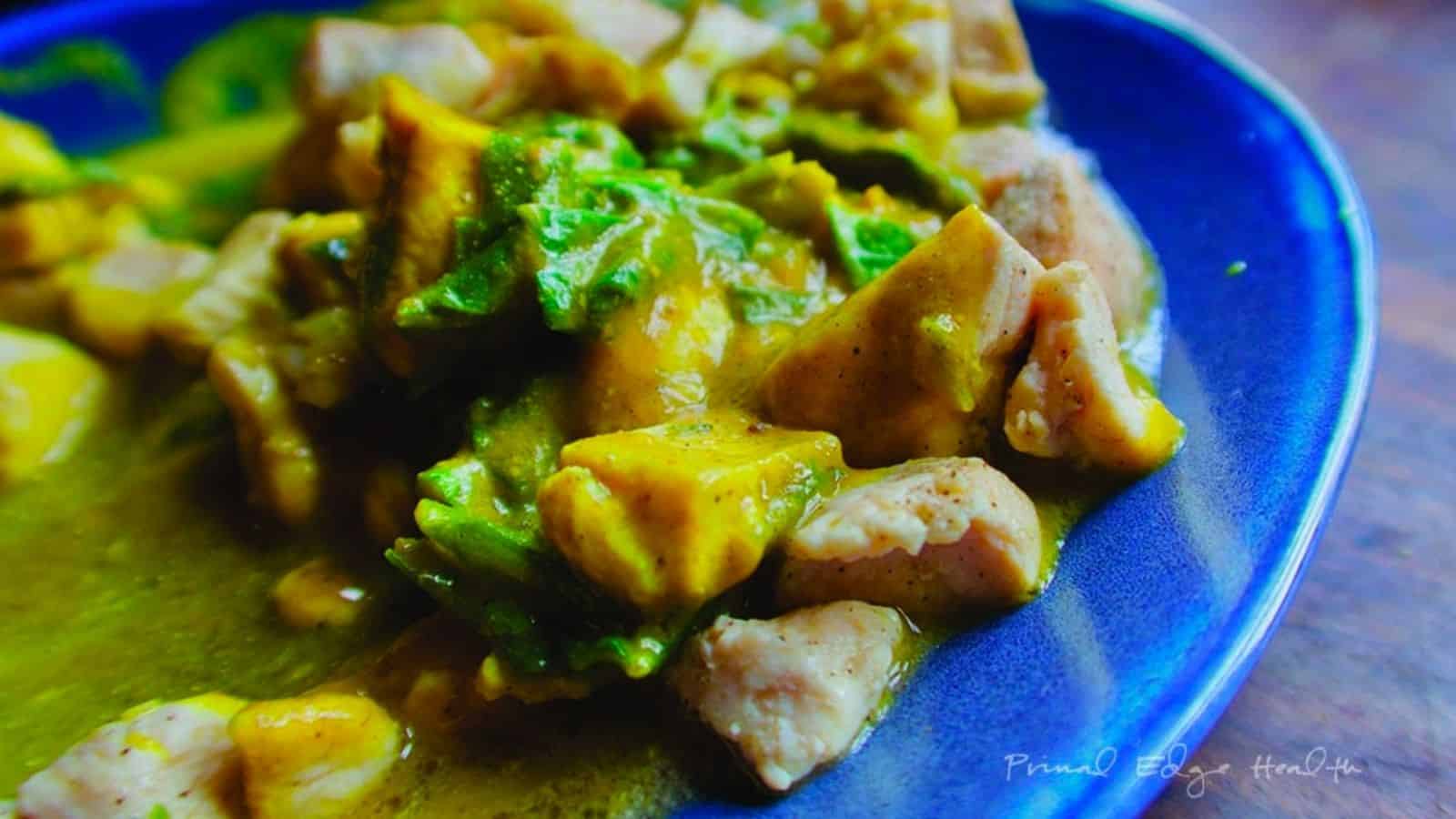 A close-up of a blue plate holding a stew with chunks of chicken, avocado pieces, and leafy greens in a yellow sauce.