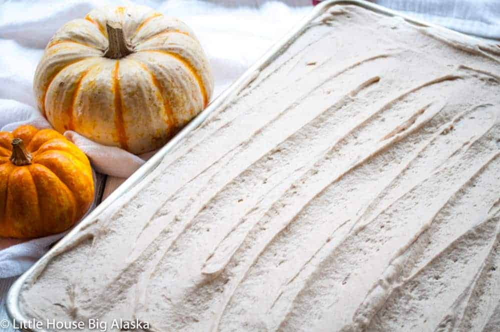 Two pumpkins sit next to a baking sheet covered with smoothed, light brown batter, likely pumpkin-flavored, on a white surface.