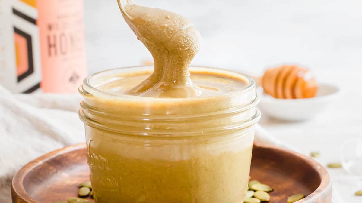 A spoon lifts creamy honey from a glass jar placed on a wooden plate, with a small bowl and honey dipper in the background.