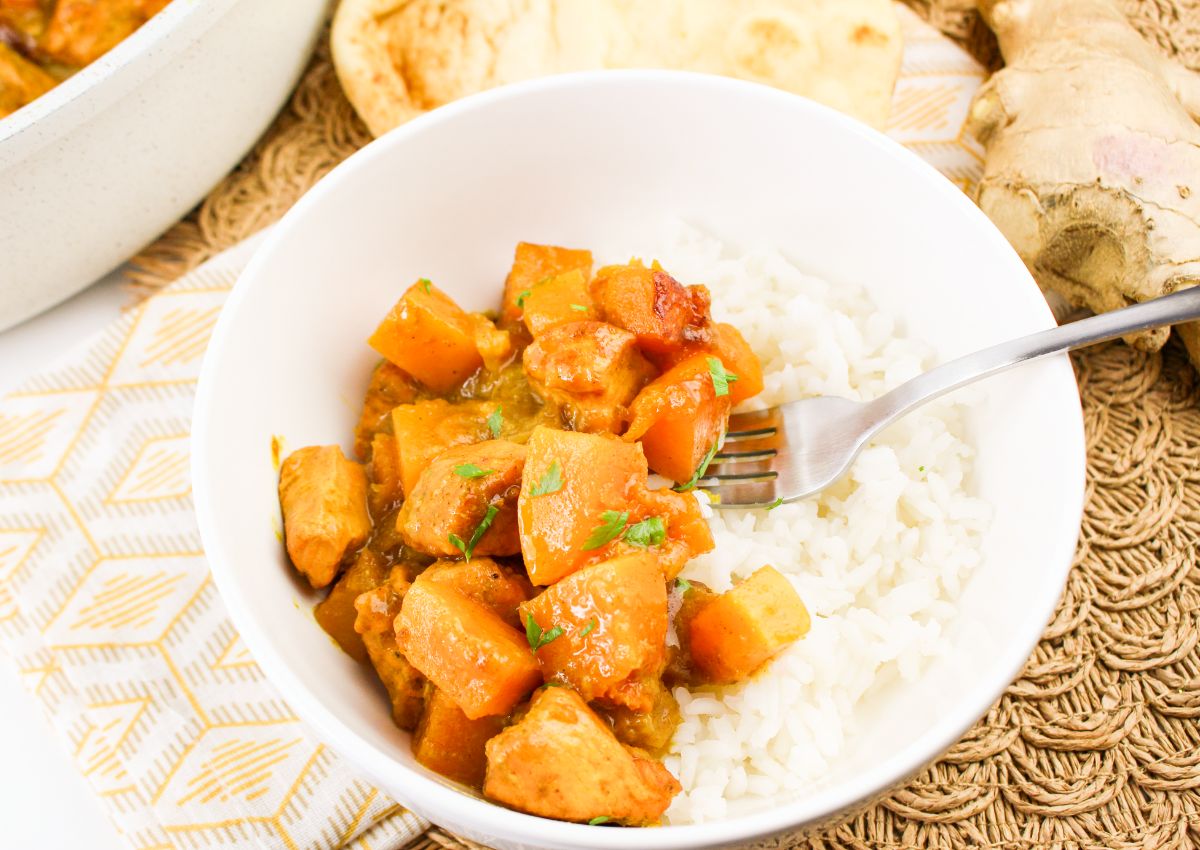 A white bowl filled with white rice and a stew containing chicken and chunks of orange vegetables, with a fork resting on the side.