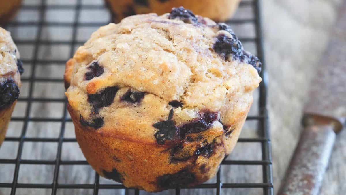 A blueberry muffin sits on a cooling rack next to a butter knife.