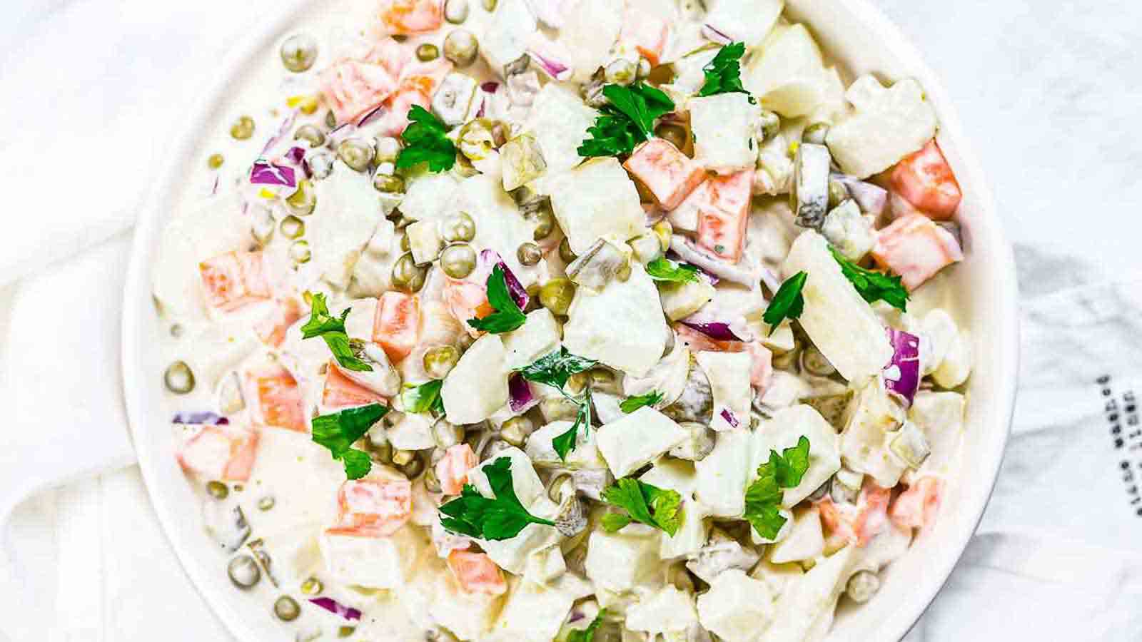 A bowl of creamy potato salad with diced potatoes, carrots, peas, red onions, pickles, and parsley garnished on top.